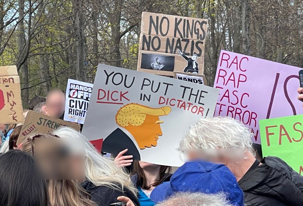 A protester holds a sign showing a cartoon of Donald Trump’s profile, his hair shaped like a dick, with the text “YOU PUT THE DICK IN DICTATOR!” Other signs nearby include “NO KINGS NO NAZIS,” “HANDS OFF CIVIL RIGHTS,” and “RESIST FASCISM.”
