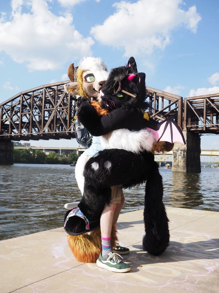 A photo of 2 fursuiters, Blanc the bison holding up Darkstar the demon cat at the waterfront. 