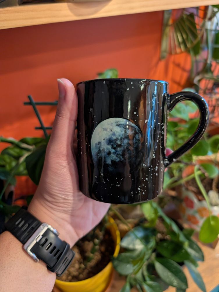 A hand holds an intricately glazed ceramic full moon mug. The background is an orange wall and house plants. 