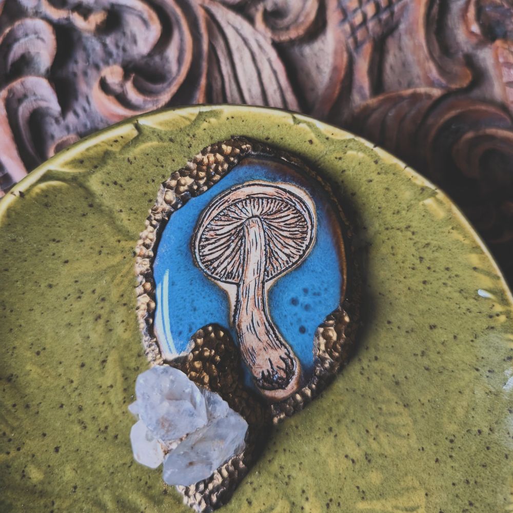 Detail shot of a green ceramic dish with a mushroom engraving on a blue tile and a mounted quartz crystal. 