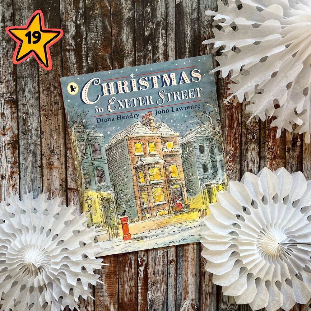 A photograph of the book cover Christmas in Exeter Street. The cover shows an illustration of a row of houses, outside on the streets there are lit street lamps, a red post box, and snow on the ground. It is snowing. The middle house shows all the windows lit up, and Santa Claus standing outside the front door.