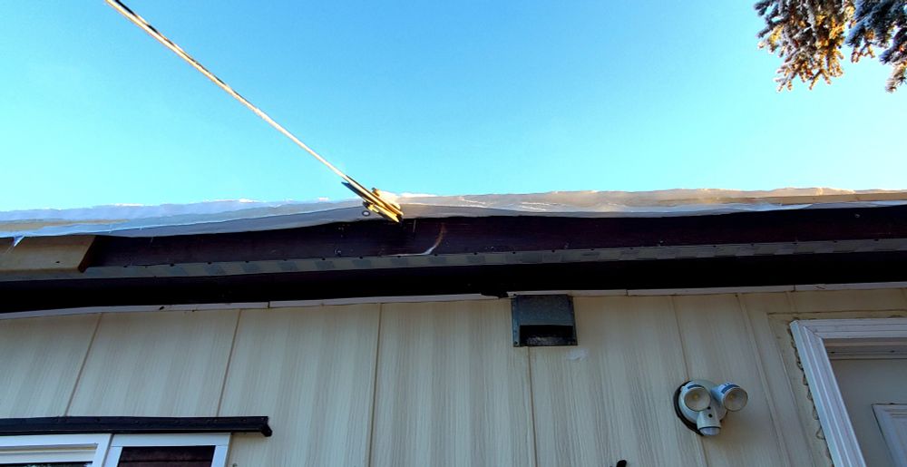 The side and roof of a beige trailer. The corners of window and door are in the left and right lower corners of the pic, respectively. A small motion sensor light is beside the door, and a vent in between, right at the roofline, and above that, a clothesline attached to the worn building.

The clothesline pushes up the edge of a white tarp serving as roofing, which is held down by a sold two inches of ice, topped with a couple more inches of snow.