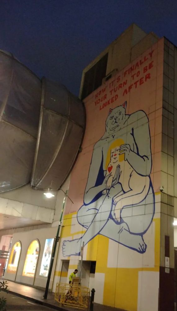 a photo of a huge artwork on the side of a tall building. there is a weirdly anthropomorphised cat petting a cat with a human face, long blonde hair, and human skin coloured fur. above it is written "Now it's finally your turn to be looked after" 