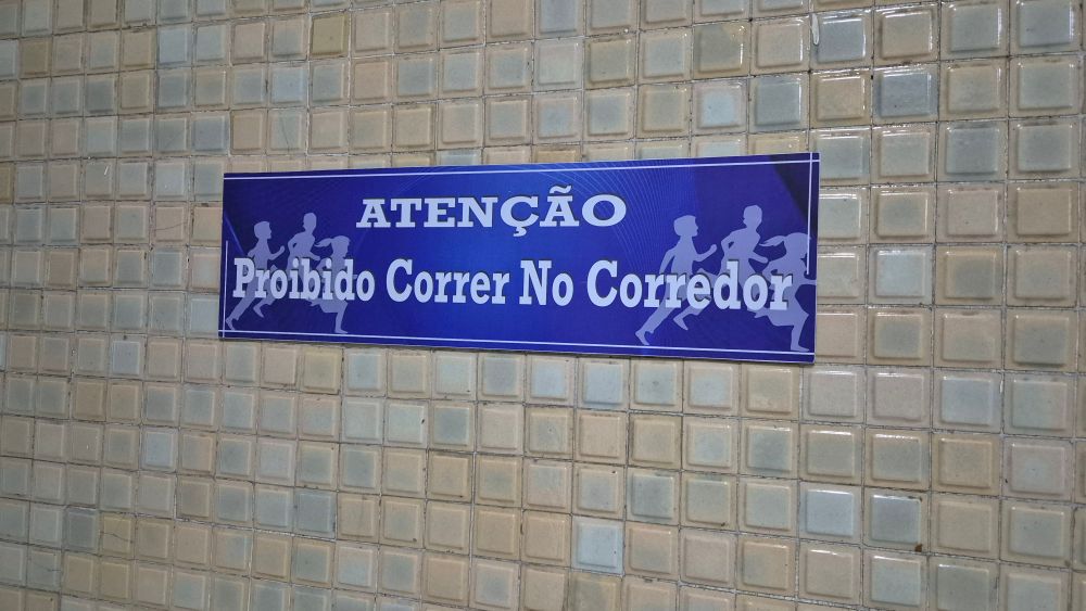 Foto de uma placa azul colada em uma parede de ladrilhos beges, com os dizeres: 

"ATENÇÃO
Proibido Correr No Corredor"

Nos cantos da placa existem desenhos de silhuetas de jovens correndo.