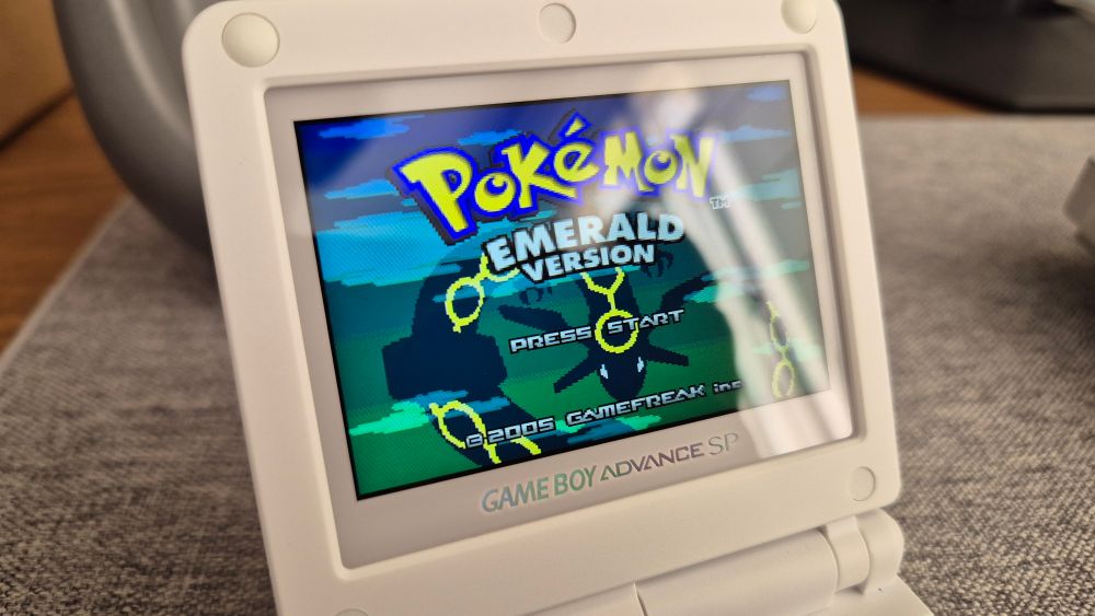 Foto da tela de um Game Boy Advance SP modificado branco mostrando a tela inicial do jogo Pokémon Emerald. A tela contém o logo do jogo, a silhueta do Pokémon dragão Rayquaza e o texto de "Press Start". Ao fundo, uma mesa de. madeira com tapete em tecido cinza