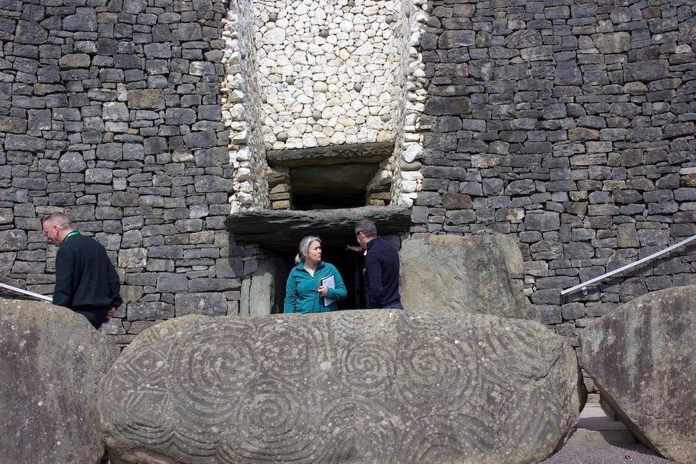 A photo of writer Caroline Pignat at Newgrange, Ireland. She has her grey hair pulled back into a ponytail and is seen wearing a turquoise zip jacket in the centre of the frame, talking to a man. In the foreground is a massive stone, etched with intricate spirals. In the background are tall stone-bricked walls, and dark entrances to places unknown.