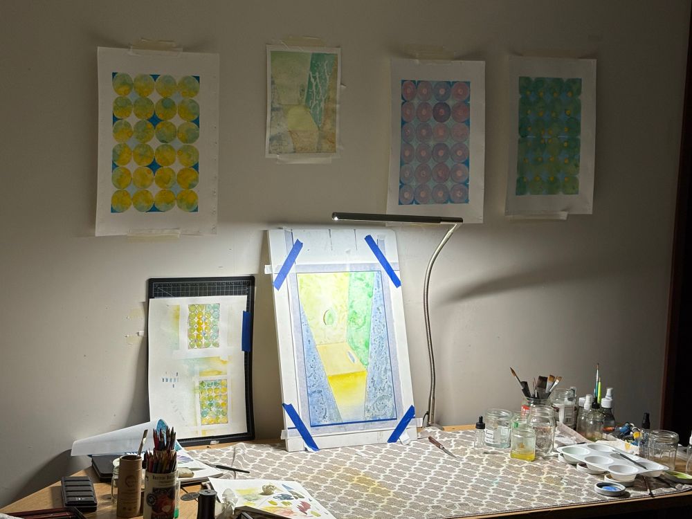 A work table cluttered with the tools for working on paper and a wall covered in various watercolor paintings. 