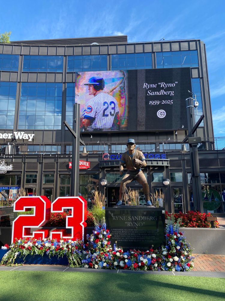 The Ryne Sandberg statue at Gallagher Way with a huge red 23 and loads of red, white and blue flowers under a large video screen with his photo and the words “Ryne ‘Ryno’ Sandberg 1959-2025”