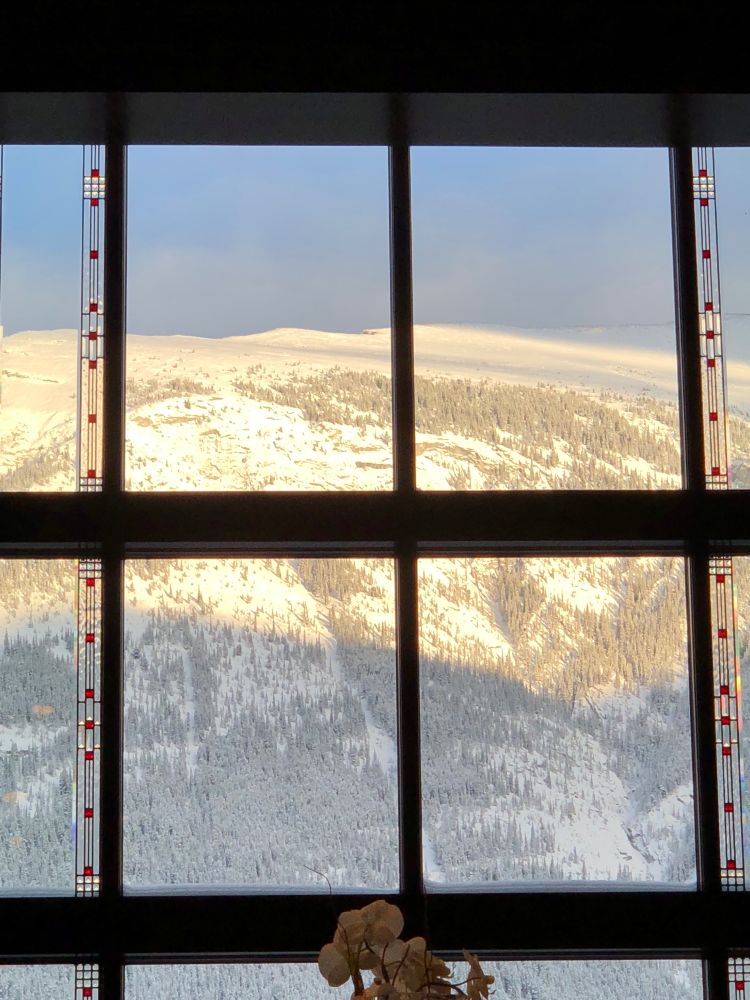 Looking through a huge paned window of the lobby of the Fairmont Banff Springs hotel onto a sunlit and snow-covered mountain under clear light blue skies. A prairie style thin vertical line of stained glass - vertical lines interspersed with small red squares - runs along the outer panes. At the bottom of the window, the light pink flowers of an orchid peek up.