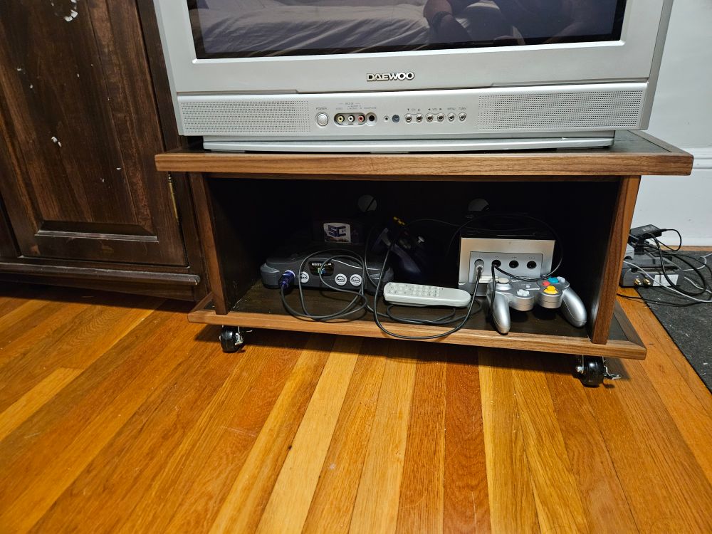 A short walnut and plywood TV stand on casters. There is a game cube and a Nintendo 64 inside the open cabinet.
