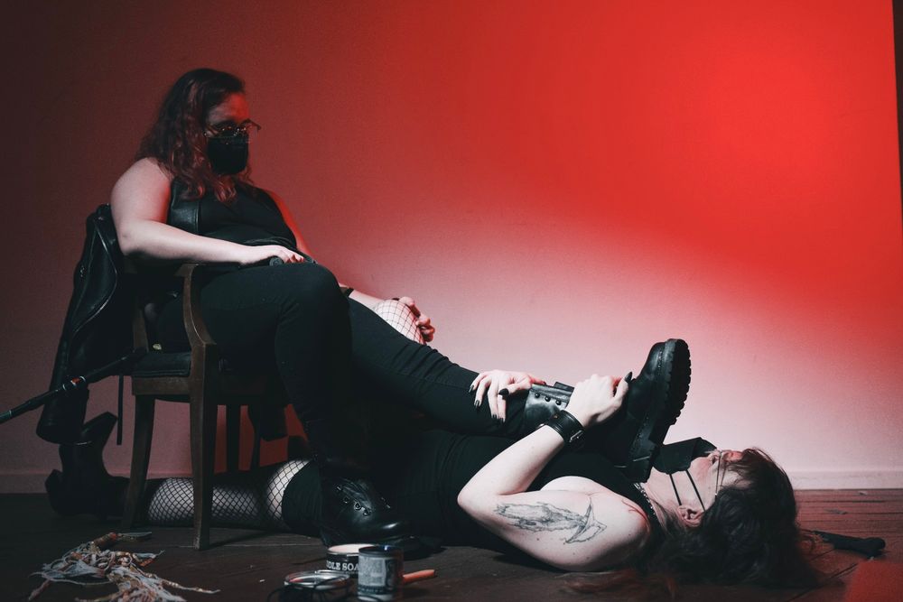 Val is sitting in a chair with Chastity lying on her back, feet underneath the chair. Val is sitting back with her feet up, one leather boot pressed under Chastity's chin. Both are dressed in all black with a lot of leather, wearing KN95 masks. The shot is from a low angle, with lots of shadows and a red background. There is leather gear and bootblack equipment strewn around the floor.