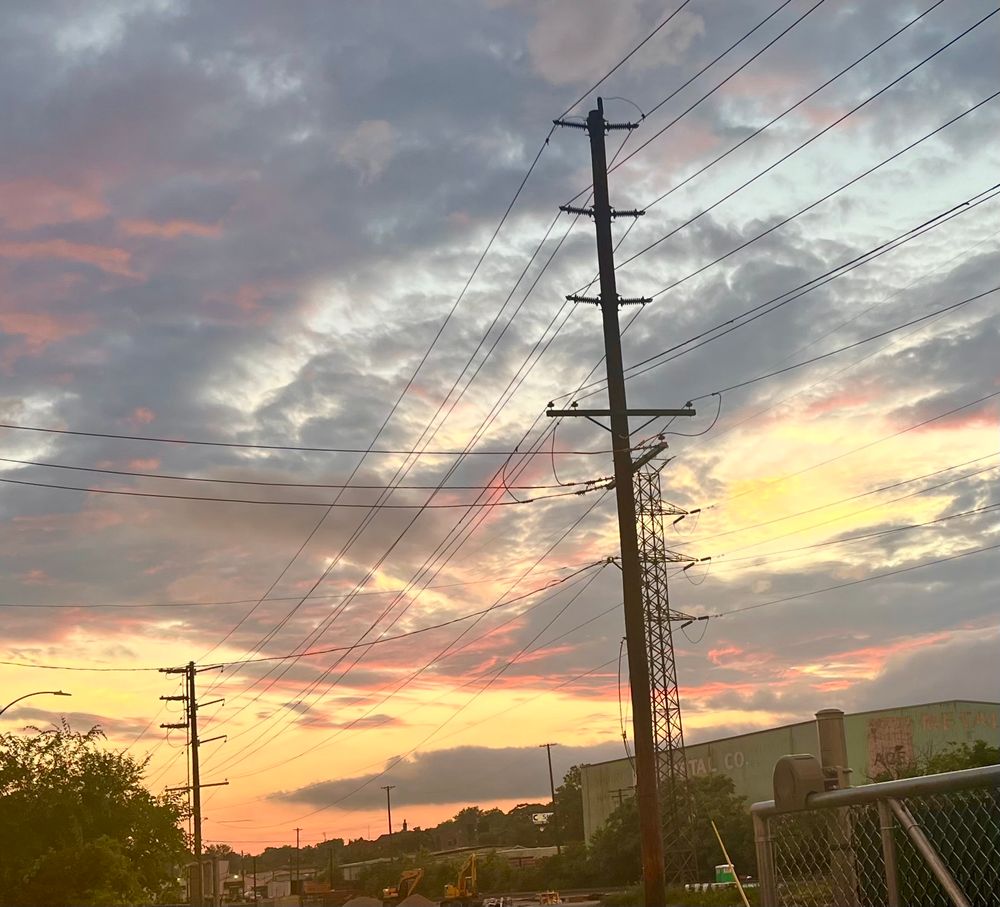 A sunset with lots of clouds reflecting warm colors on their bases, tree line at the very bottom with a telephone pole near right side. 