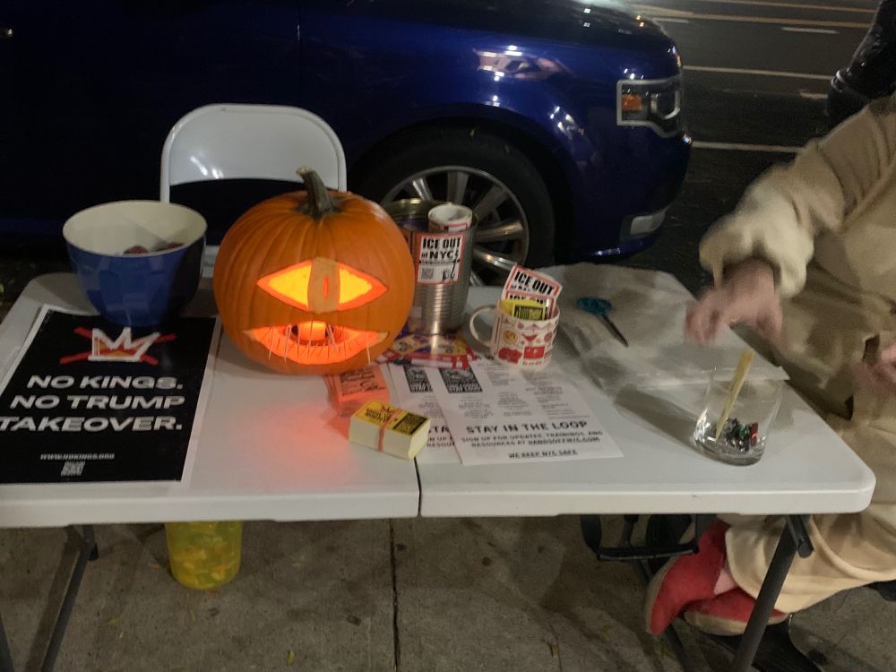 A folding table on a city sidewalk is covered with a halloween jack-o-lantern, bowls of chocolates and baby potatoes, political fliers, rolls of stickers, and a jar of miniature whistles.