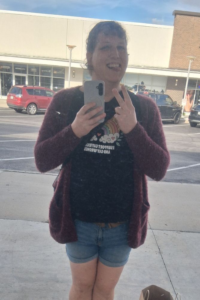 A mirror selfie in an opaque window. I'm standing, flashing a peace sign, smiling and sticking out my tongue (I haven't figured out smiling cute for pictures without that last step; this is still new to me). My hair is back in a ponytail. I'm wearing a purple(?) fuzzy cardigan, a black tee which reads (albeit mirrored), "I Support Gay Rights and Gay Wrongs," and denim shorts