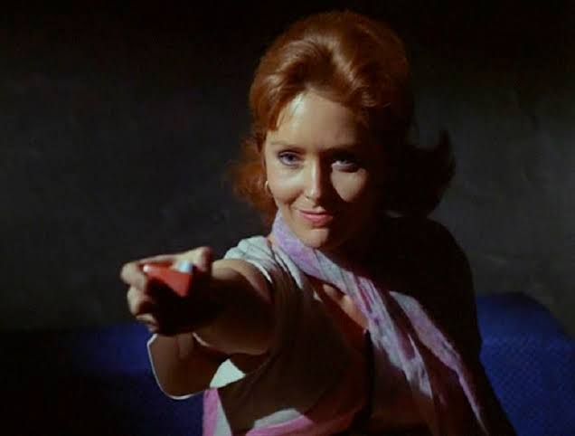 A redheaded woman in a reclining position holds out an item of unclear nature as if it's a weapon and she is tauntingly making demands.