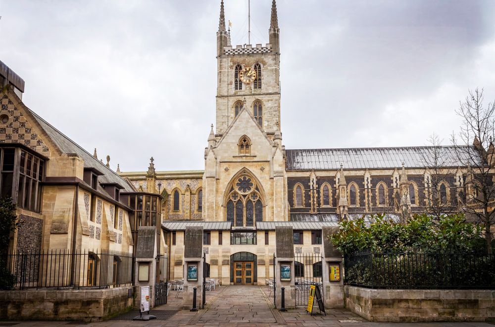 Southwark Cathedral, captured in a wide-angle shot, showcases its intricate Gothic architecture and surrounding structures as it stands against the cloudy sky.