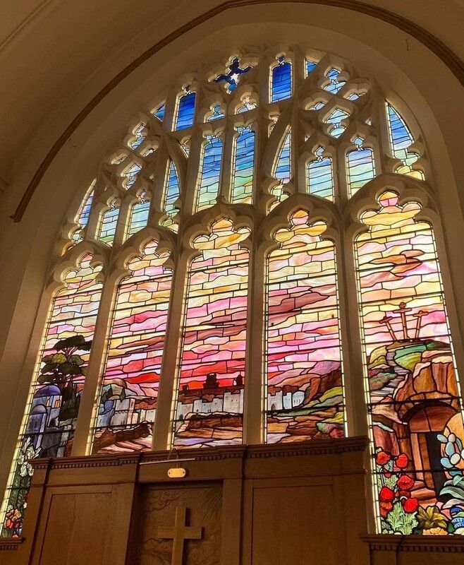Multicoloured stained glass window at St John's Church, Ainsdale, depicting the city of Jerusalem, three crosses on a hill outside the city and Jesus' tomb with stone rolled away.