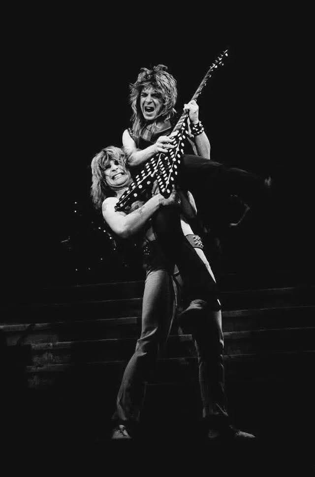Photograph of Ozzy Osbourne lifting up Randy Rhoads who is playing a guitar.