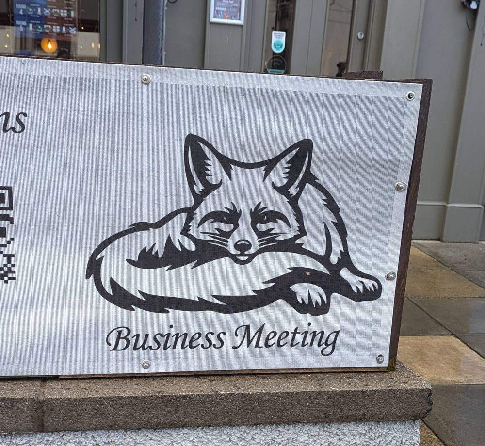 Photo of a sign outside a venue. The sign has a grey background and a black image of a fox resting its chin on its tail. The sign reads "Business Meeting" below the fox