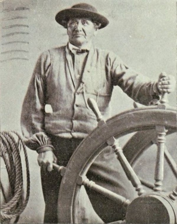 A man holds a ship's wheel with both hands.  He is quite tall. 