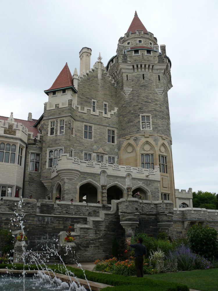 The main turret of Casa Loma, a gray brick castle, rises against the sky. There is a fountain in the foreground, and a stone wall.