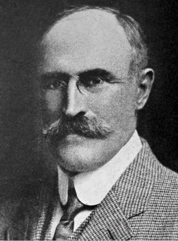 A black and white image of a balding white man with small glasses and a bushy mustache. He is wearing a suit and tie, his white shirt has a large collar. 