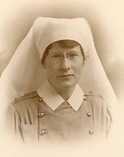 A white woman is wearing a white nurse's headdress that flows behind her shoulders. Her outfit has military-style buttons and a large white collar. She is wearing glasses and is looking into the camera. 