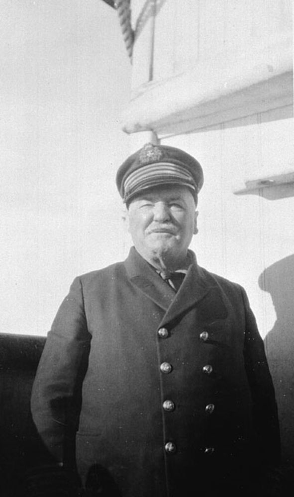 A black and white image of a man in a naval-style jacket and hat who squints into the sun.