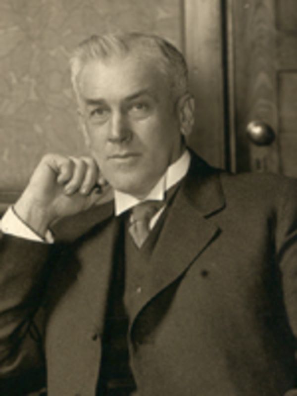 Sepia-toned photo of a white man wearing a suit and tie. His hair looks gray/silver and his eyebrows are darker. 