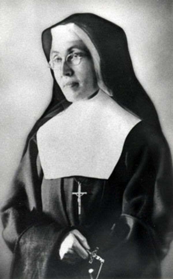Black and white photo of a nun, wearing a traditional black and white habit and a large cross around her neck. She is wearing glasses and her hair is covered.