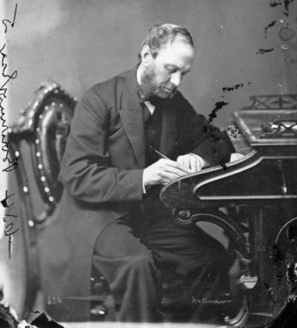 A white man with a high forehead and thick sideburns is wearing a dark suit. He is sitting in a chair, looking down at the paper he is writing on which is balanced on an elaborate desk.