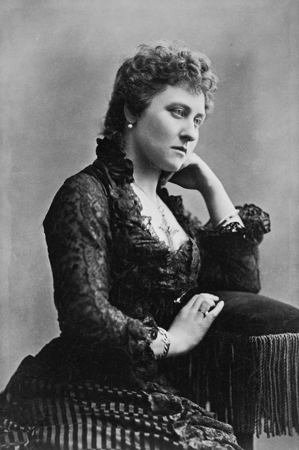 A white woman with curly hair wears a dress with a ruffled black top and a vertically striped skirt. She leans on a pillow with long fringe, and looks off camera. She is wearing a large ornate cross on a chain around her neck.