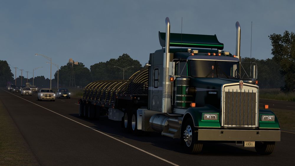 The Kenworth W900 from the front, driving on a two-lane road with grass-lined shoulder. Trees line both sides of the road, with many streetlights and power lines. The morning sky is soft pastel blue, and traffic is moderate, with many vehicles on the road.
