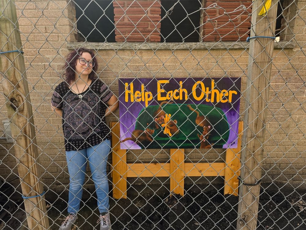 Art mural is against a brick wall, the artist is standing next to it and they're behind a chain-link fence 