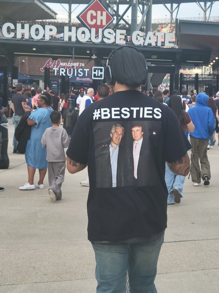 picture of the back of a man in front of truist park in Atlanta. the back of his shirt has a picture of donald trump and jeffery epstein and is captioned #besties