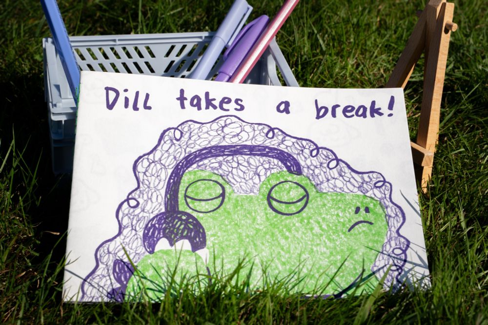 Photograph of my picture book called "Dill takes a break!" in an A5 horizontal format. The cover shows a vibrant green textured crocodile character putting on purple headphones with a decisive look on their face. It's my second picture book, printed with riso.
The book is standing on grass, leaning of a box with pens.