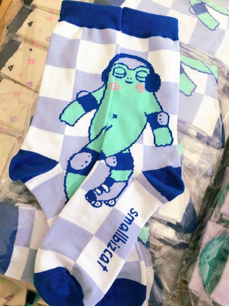 Picture of a pair of socks laid out so you can see the whole design. It shows a rollerskating frog all geared out with head phones and protectors. The outlines, cuff, heel and toes are a rich blue color, same for the name on the sole "smallbizcat", the background a lavender and white checkerboard. The frog is green with big swirly pink cheeks, he looks very content 😌