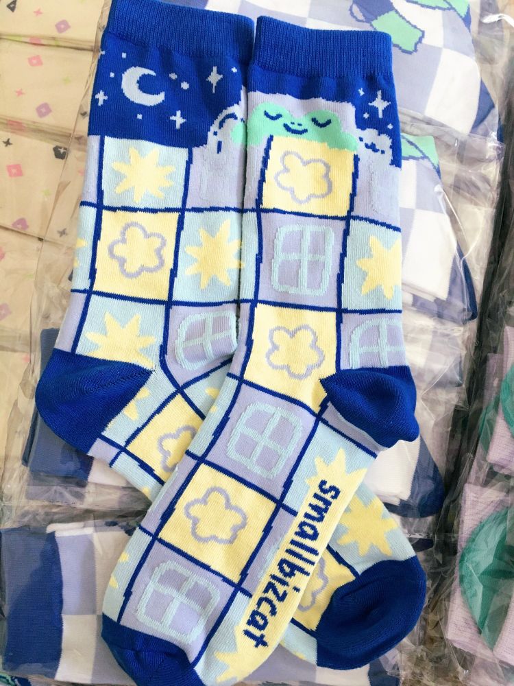 Picture of a pair of socks laid out so you can see the whole design. It shows mostly a patchwork patterened blanket all over the sock with a little frog sticking out at the top laying on a pillow. Next to him are a shark and a bear plushie also tucked in. Behind them the night sky. The blanket pattern includes light blue parts with yellow stars, yellow parts with purple flowers and purple parts with blue "windows". The outlines, cuff, heel and toes are a rich blue color, same as the name on the sole "smallbizcat".