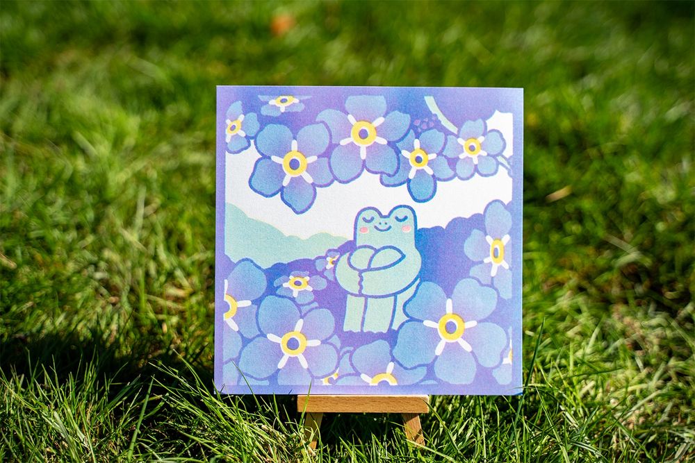 Photograph of a square risograph print of a frog chilling in a field of blue flowers. The frog is sitting down with his arms holding his legs, he looks very calm with his eyes closed and a smile on his face. The print is rested on a small standee that is standing in a field of grass.