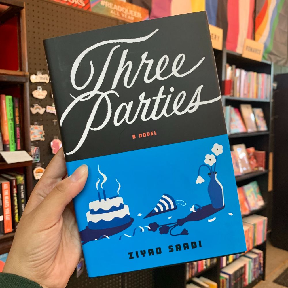 A hand holds up a copy of Three Parties by Ziyad Saadi. The top half of the cover is black with white text for the title, and the bottom half is blue and white. The bottom half of the cover shows a birthday cake, a toppled birthday party hat, and wilting flowers. 