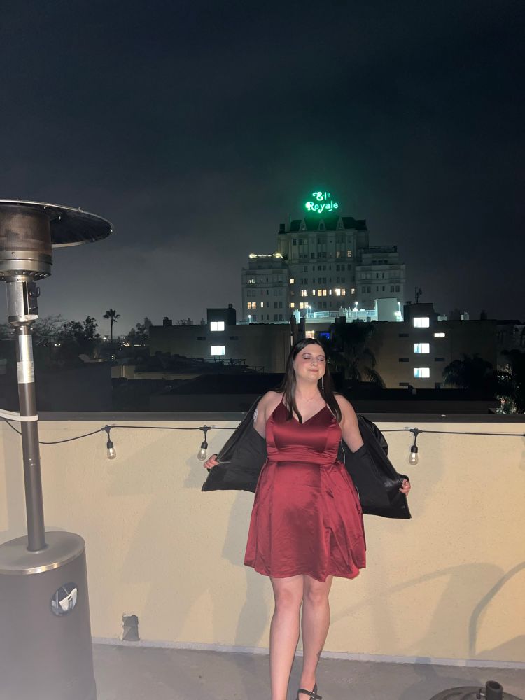 Me (a white woman in my 30s with dark brown hair and dark brown eyes wearing a shirt flared shiny red dress and leopard flats) standing on a rooftop pointing my toe and closing my eyes with a building in the background reading “el royale”