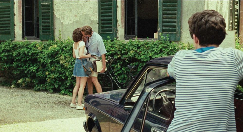 a frame from the film Call Me by Your Name. Elio leans against the car and watches Oliver kissing Chiara. 