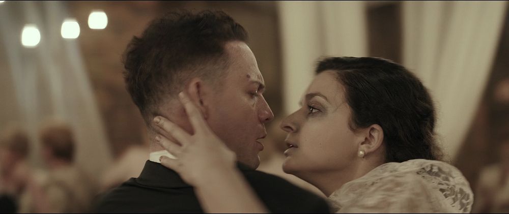 A woman in a wedding dress reaches towards the neck of a man in a suit. From the film Demon (2015).