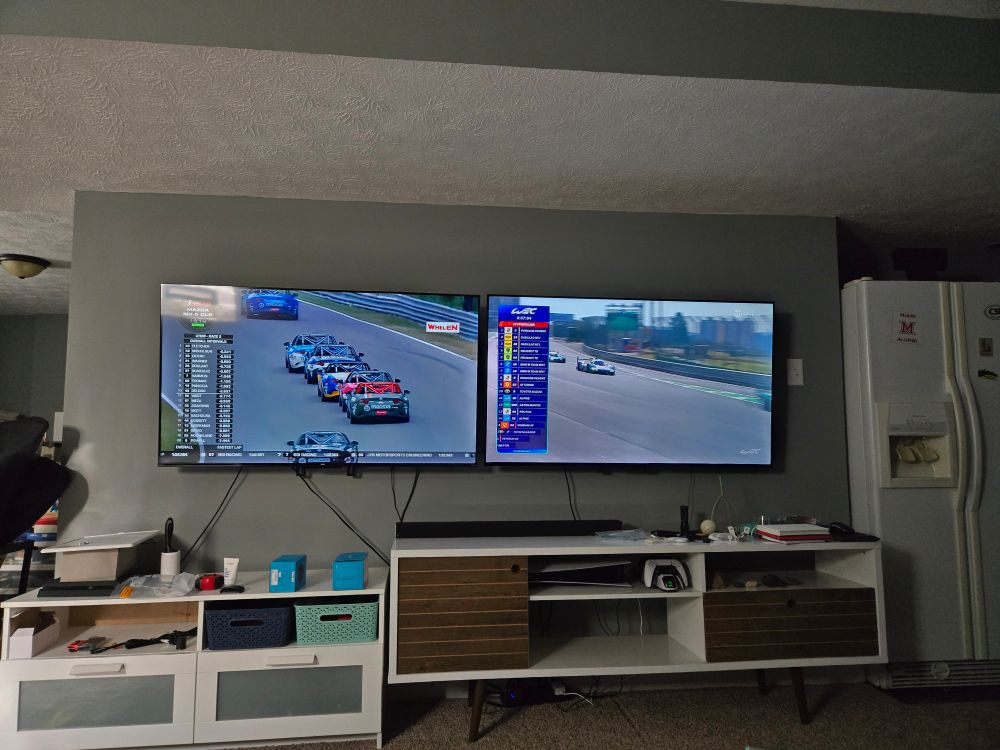 Two tvs in a basement, one showing MX5 Cup and the other with World Endurance Chapionship on it. 