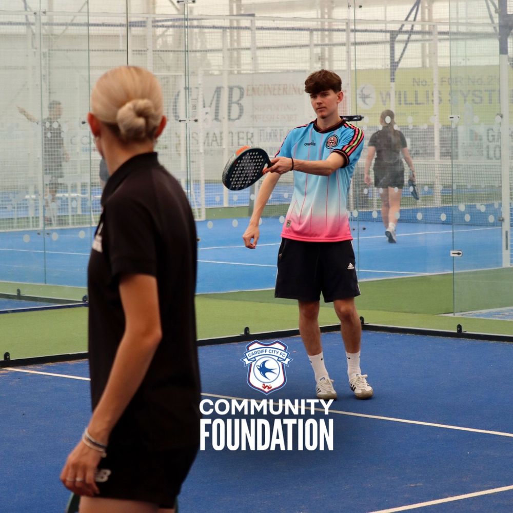 Two young people playing padel in an indoor court, one in the foreground partially visible and the other in the background holding a padel racket.