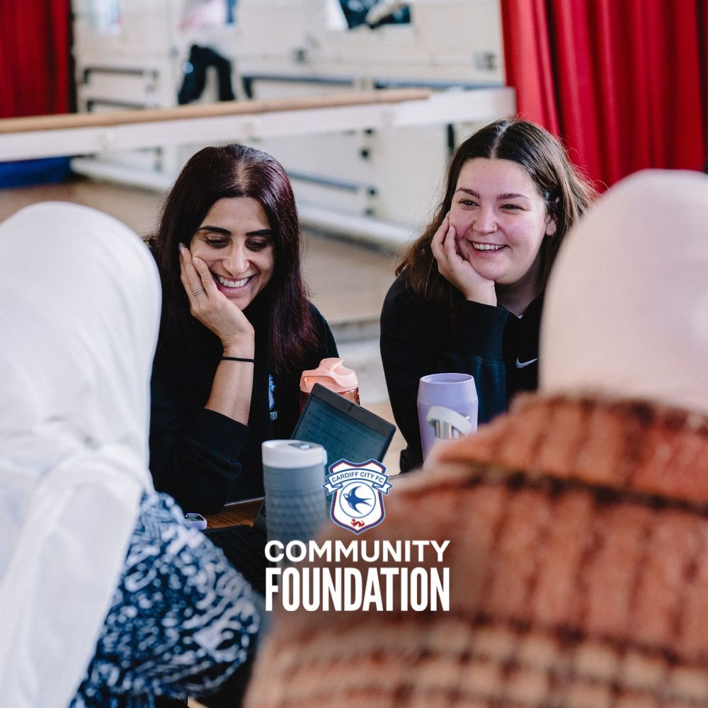 Photo by Cardiff City FC Community Foundation displaying two women smiling and sat at a table, showing a laptop screen to the rest of the female participants at the table.