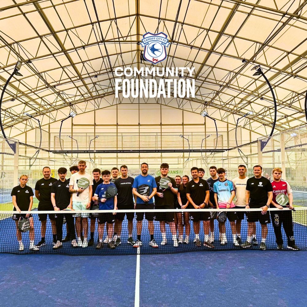 Group photo of participants and staff at an indoor paddle court, standing in front of a net, posing with paddle sticks.