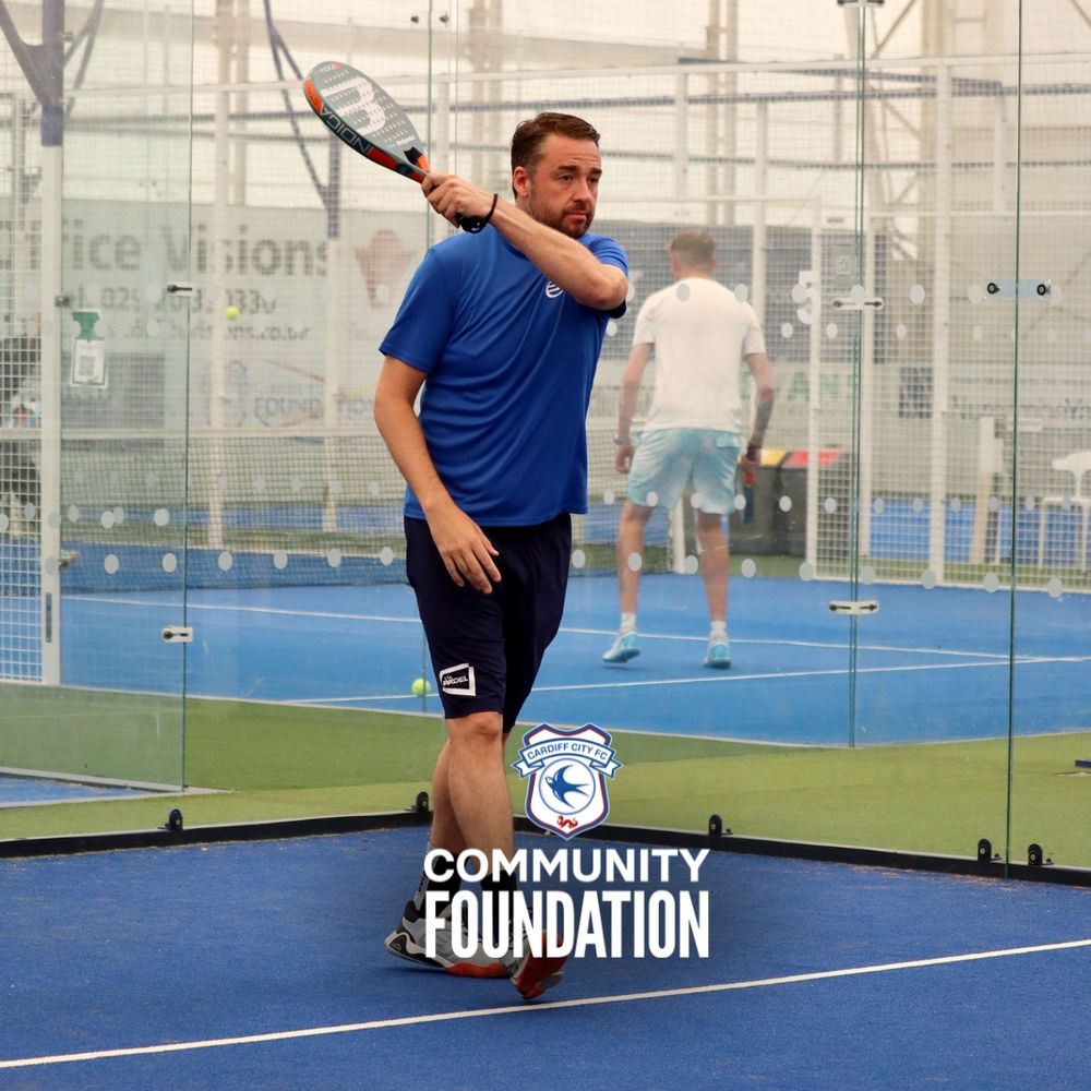 Jason Manford playing padel on an indoor court, wearing a blue shirt and shorts.