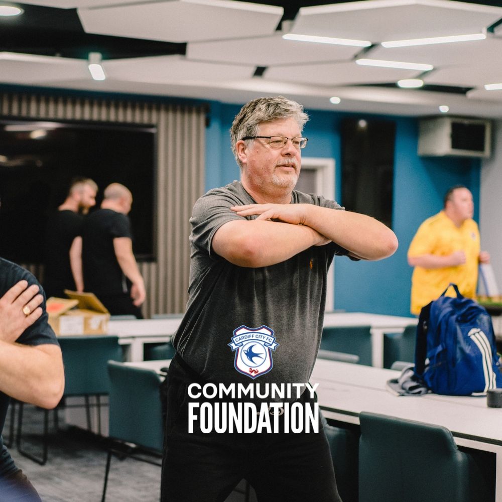 Photo by Cardiff City FC Community Foundation showing male crossing his arms and commencing to bend down during exercise move, while people walk by behind him.