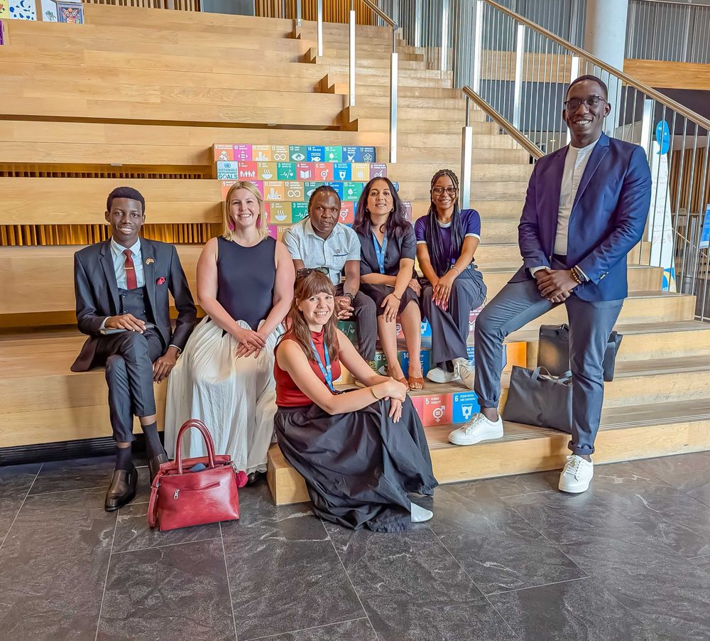 A group of 7 people in formal attire seated in stairs, or standing next to them, posing and smiling to the camera.
